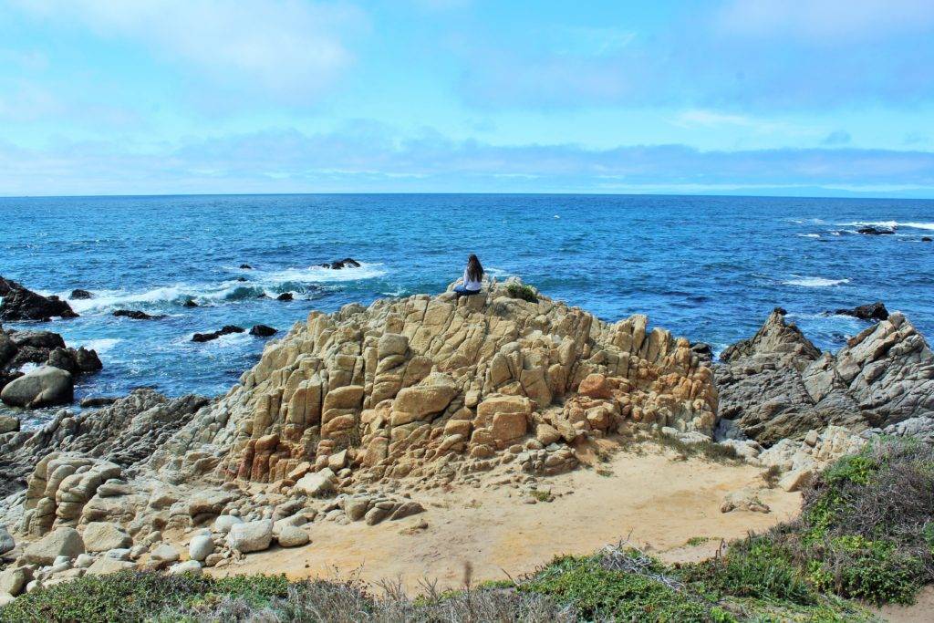 Mandala Traveler sitting on a large rock formation with the pacific ocean in the background