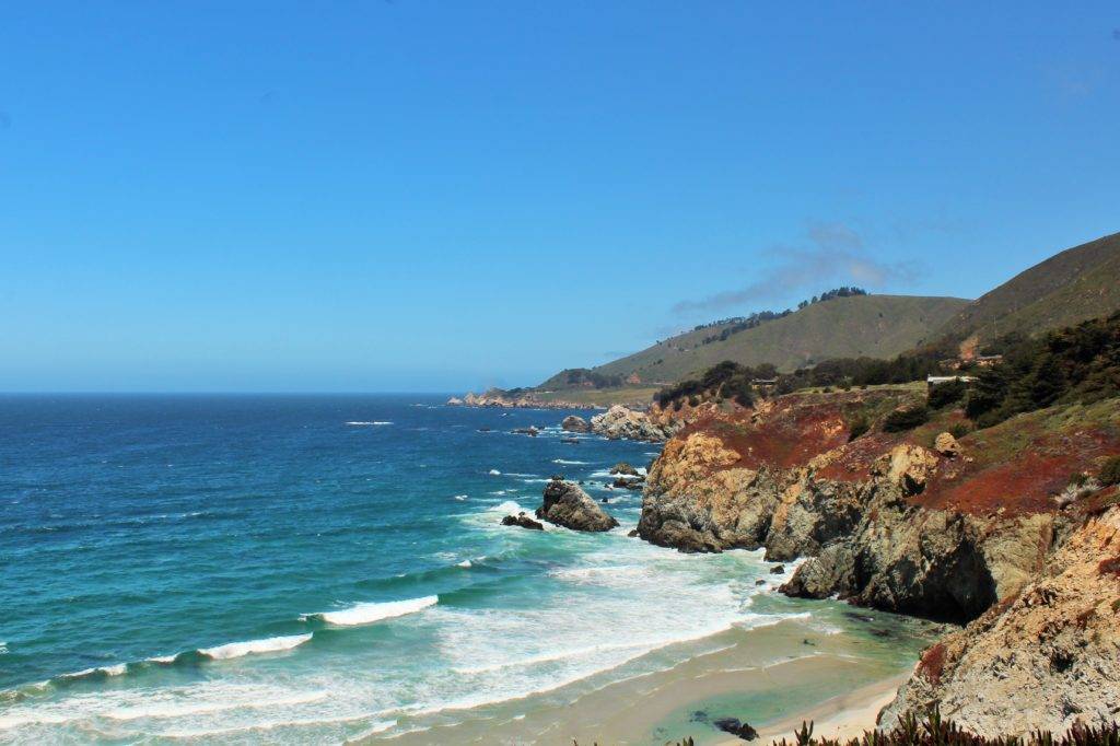 Rocky Point on Highway 1. Bright blue sky and ocean with green hills and red wildflowers on the cliffs.