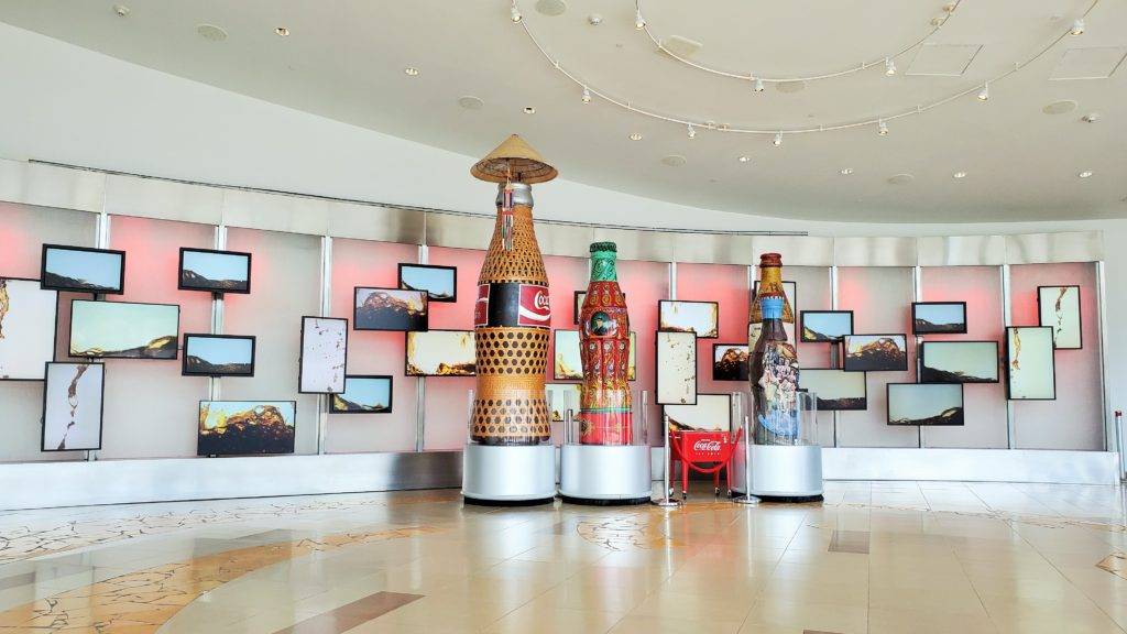 White room with many televisions featuring Coca-Cola being poured. 4 large bottles made from creative materials in the middle