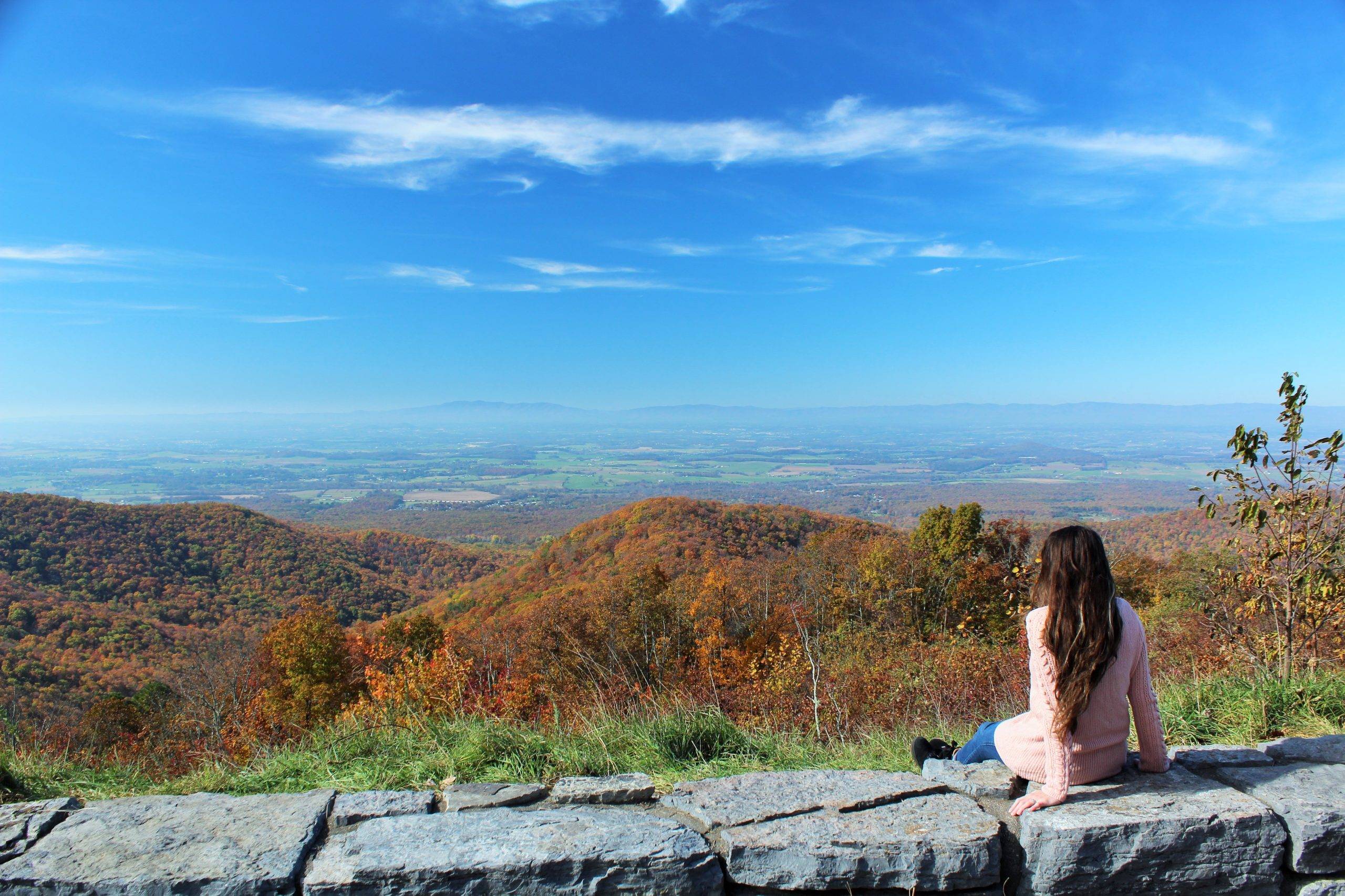 Girl in a pink sweater sitting on a low stone wall with colorful mountains and valleys in the background