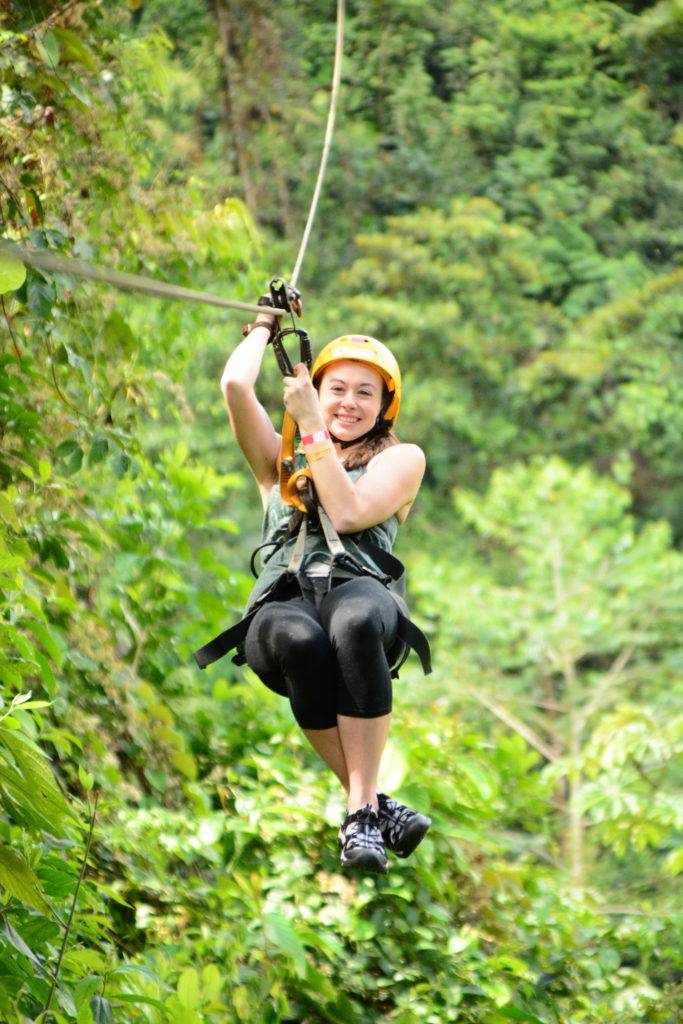 girl hanging on zip line with yellow helmet and green rainforest in the background