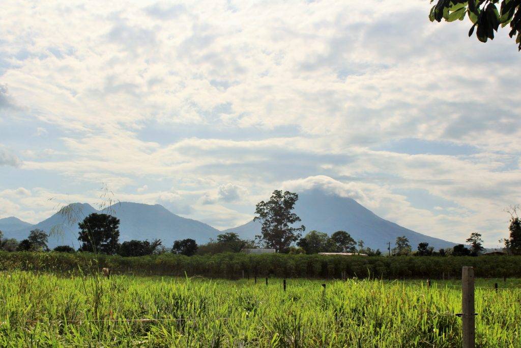 Volcano with top covered by clouds with mountains in the background and green grass and plants in the foreground