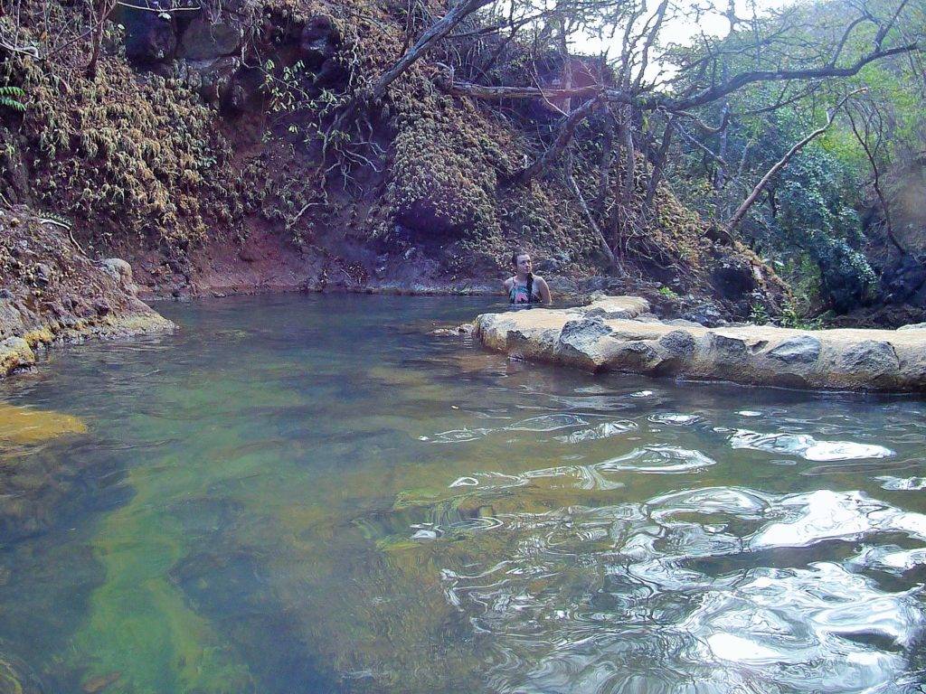 A girl in a pink swimsuit sitting in a thermal spring lined with rock and a dirt river bank