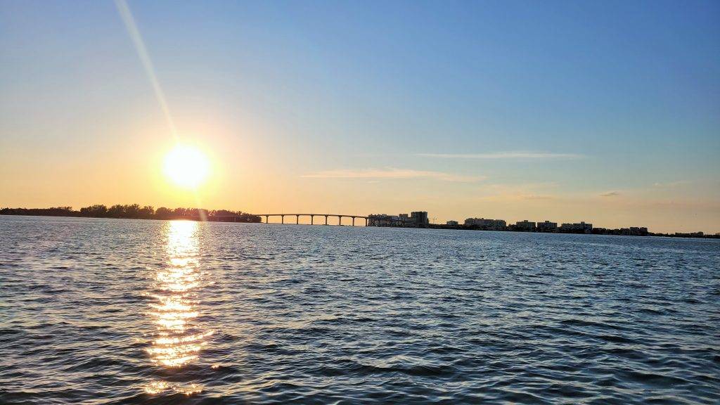 sun setting in the distance over a bridge with ocean in the foreground. 