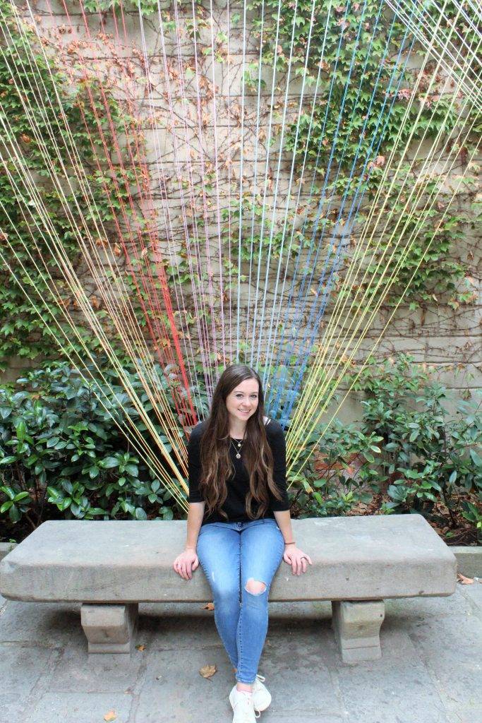 Girl sitting on a stone bench with colorful strings and an ivy-covered wall in the background