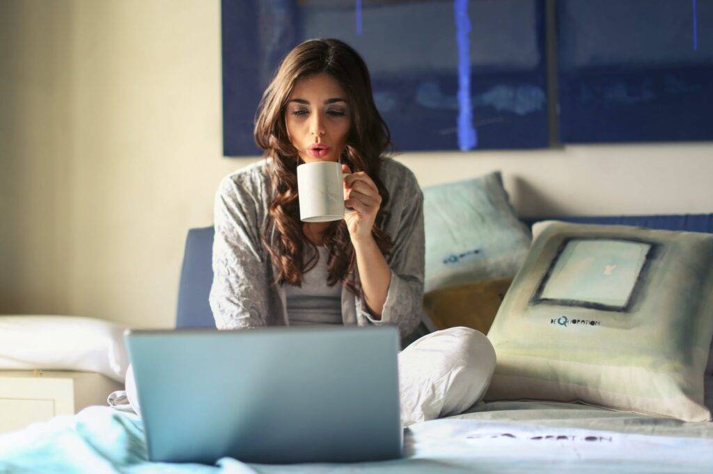 woman in grey jacket sits on bed uses grey laptop work from hotels