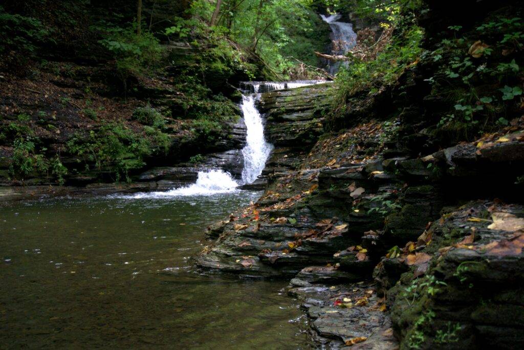 Small tiered waterfall flowing into a rock-lined river in the woods