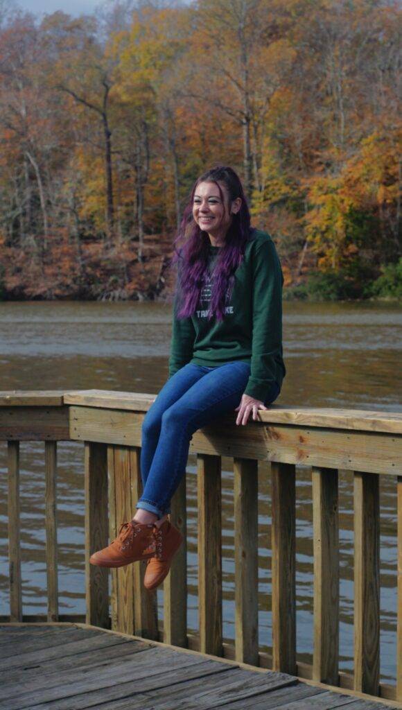 Girl wearing BANGS sneakers sitting on a fence with water and fall foliage in the background